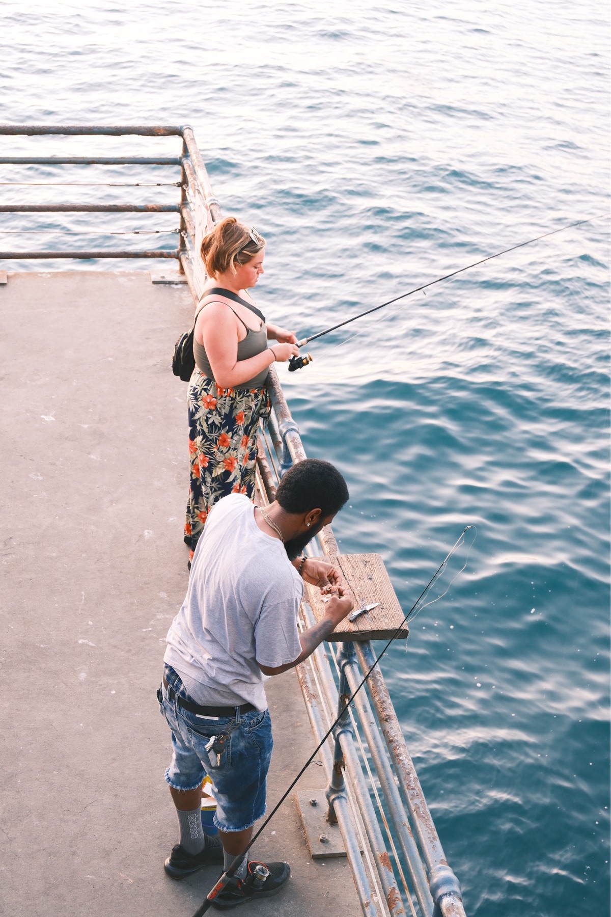 Two people preparing fishing lines from a pier above blue water